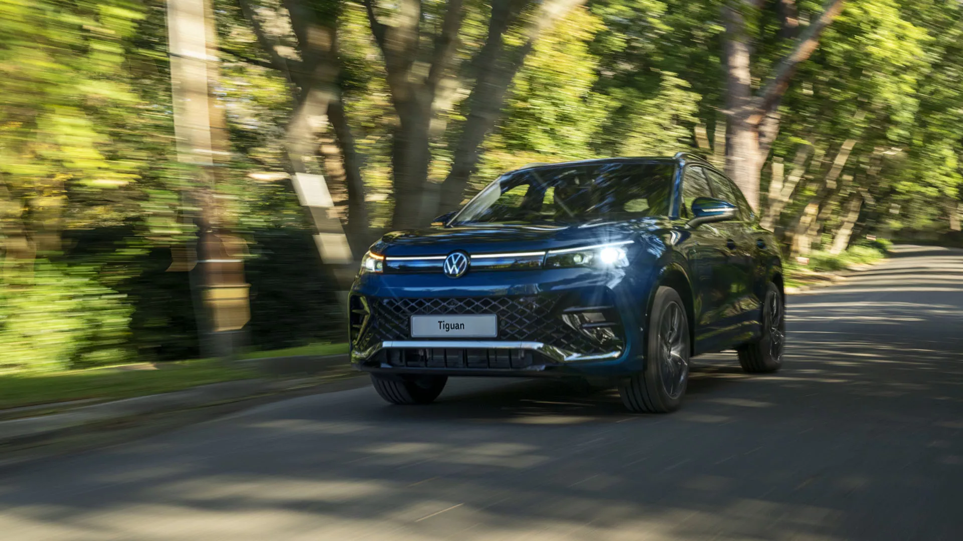 Green VW Tiguan parked in front view on the driveway of a modern apartment building. A young couple stands next to it and embraces.